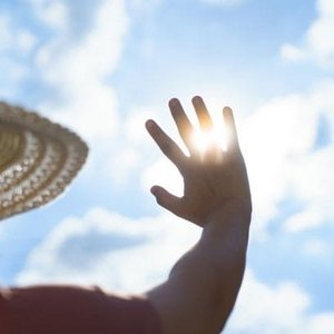 Person mit Strohhut hebt Hand gegen die Sonne, um sich vor grellem Sonnenlicht zu schützen, vor blauem Himmel mit weißen Wolken.